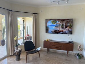 A room with a large window, a chair, a small table, a cabinet, and a painting of a coastal scene. - Beachfront condo in Paradise 3 bed /2 bath (San José del Cabo)