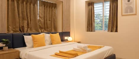 A hotel room with a bed, bedside table, air conditioning unit, window with curtains, and a wall-mounted artwork.