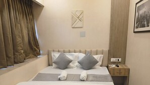 Un lit bien fait, avec des oreillers et une couverture, une table de chevet avec un téléphone et une télécommande, une lampe fixée au mur et une fenêtre avec des rideaux.