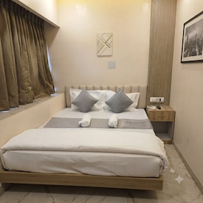 A neatly made bed with pillows and a blanket, a bedside table with a phone and remote, a wall-mounted light, and a window with curtains. - Townhouse-Executive Suites (Barasat)