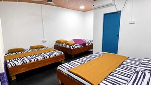 Quarto com três camas de madeira, cada uma com roupa de cama listrada e um cobertor marrom. Há uma porta azul e um aparelho de ar condicionado na parede.