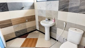 Um banheiro com piso azulejado, pia branca assentada, vaso sanitário e um desentupidor vermelho.