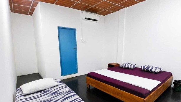 Quarto com duas camas, uma com colcha listrada e outra com colcha lisa. Há uma porta azul e uma pequena mesa com abajur.