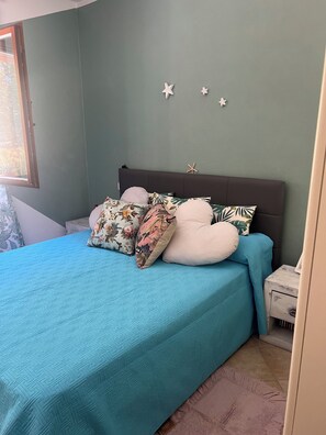A bed with a teal bedspread and multiple pillows, a wooden nightstand with a lamp, and a starfish decoration on the wall. - House with a garden in Santa Teresa di Gallura (Santa Teresa di Gallura)