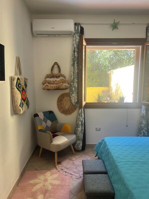 Una habitación con cama, una silla, aire acondicionado de pared, ventana con cortinas y adornos en la pared.