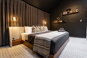 A modern bedroom with a large bed, dark curtains, wall-mounted lights, and a shelf with framed pictures. - Montrose Luxury Stay | Prime Location + Patio (Houston)