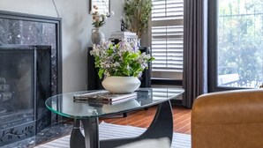 Ein Wohnzimmer mit einem Glastisch, einem Kamin, einer Topfpflanze und Blick auf Bäume durch das Fenster.