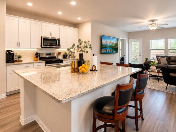Una cocina moderna con gabinetes blancos, una isla grande con taburetes de bar, un microondas y un comedor con sofá y sillas.
