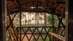 A wooden structure with a thatched roof, a bamboo fence, and a view of a beach with palm trees. - Sukumara Homestay Unique Eco Luxury Mud and Bamboo Beachfront Villa (Thrissur)