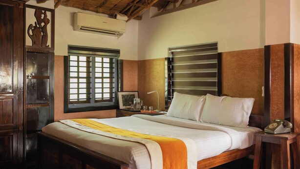 A hotel room with a wooden bed, a nightstand with a lamp, a window with bars, and a wall-mounted air conditioner. - Sukumara Homestay Unique Eco Luxury Mud and Bamboo Beachfront Villa (Thrissur)