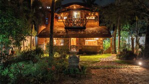 A two-story wooden house with a balcony, surrounded by lush vegetation and a stone pathway. - Sukumara Homestay Unique Eco Luxury Mud and Bamboo Beachfront Villa (Thrissur)