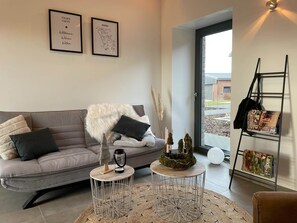 A modern living room with a grey sofa, two small round tables, a coffee table with decorative items, and a bookshelf with books and a ladder. - Lindenglück Vacation Rental (Uedem)