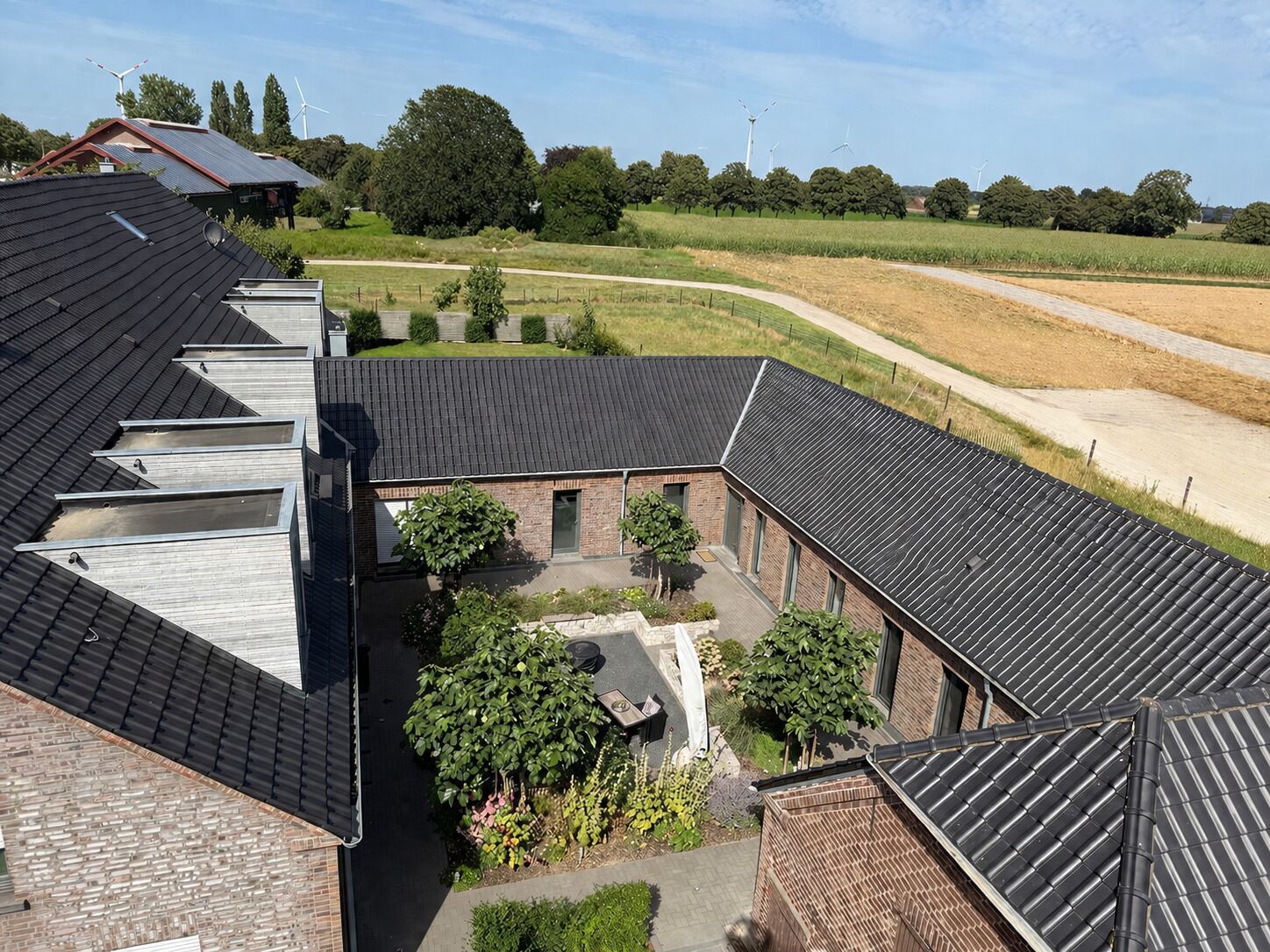 A building with multiple roof sections, a courtyard with trees, and a rural landscape with wind turbines in the background.