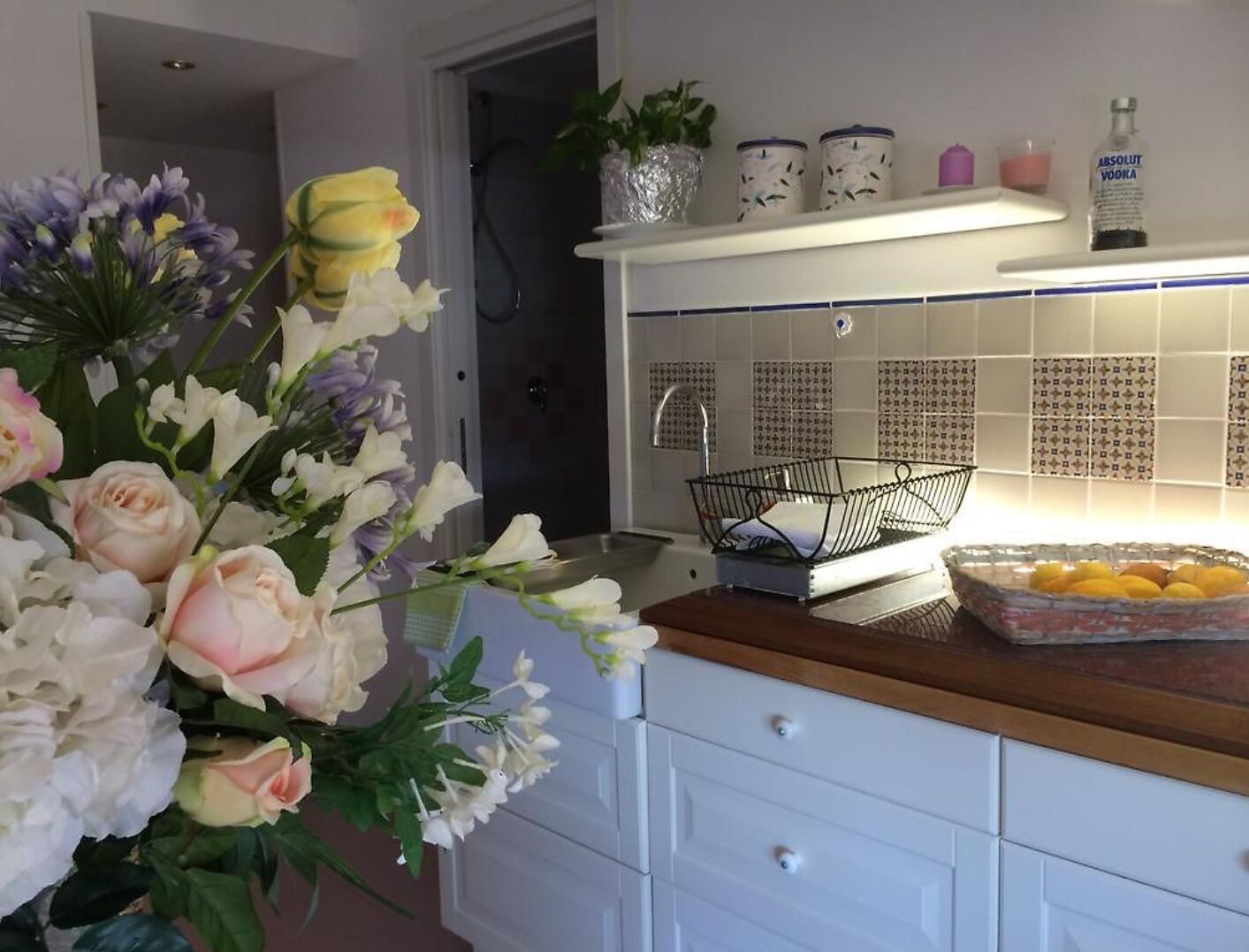 Una cucina con mobili bianchi, un piano di lavoro in legno, un vaso con fiori e uno scaffale con utensili da cucina.