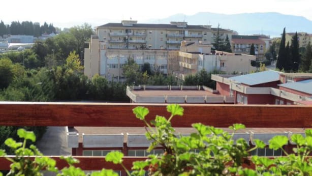 Un edificio de varios pisos con techo plano, rodeado de vegetación y otros edificios.
