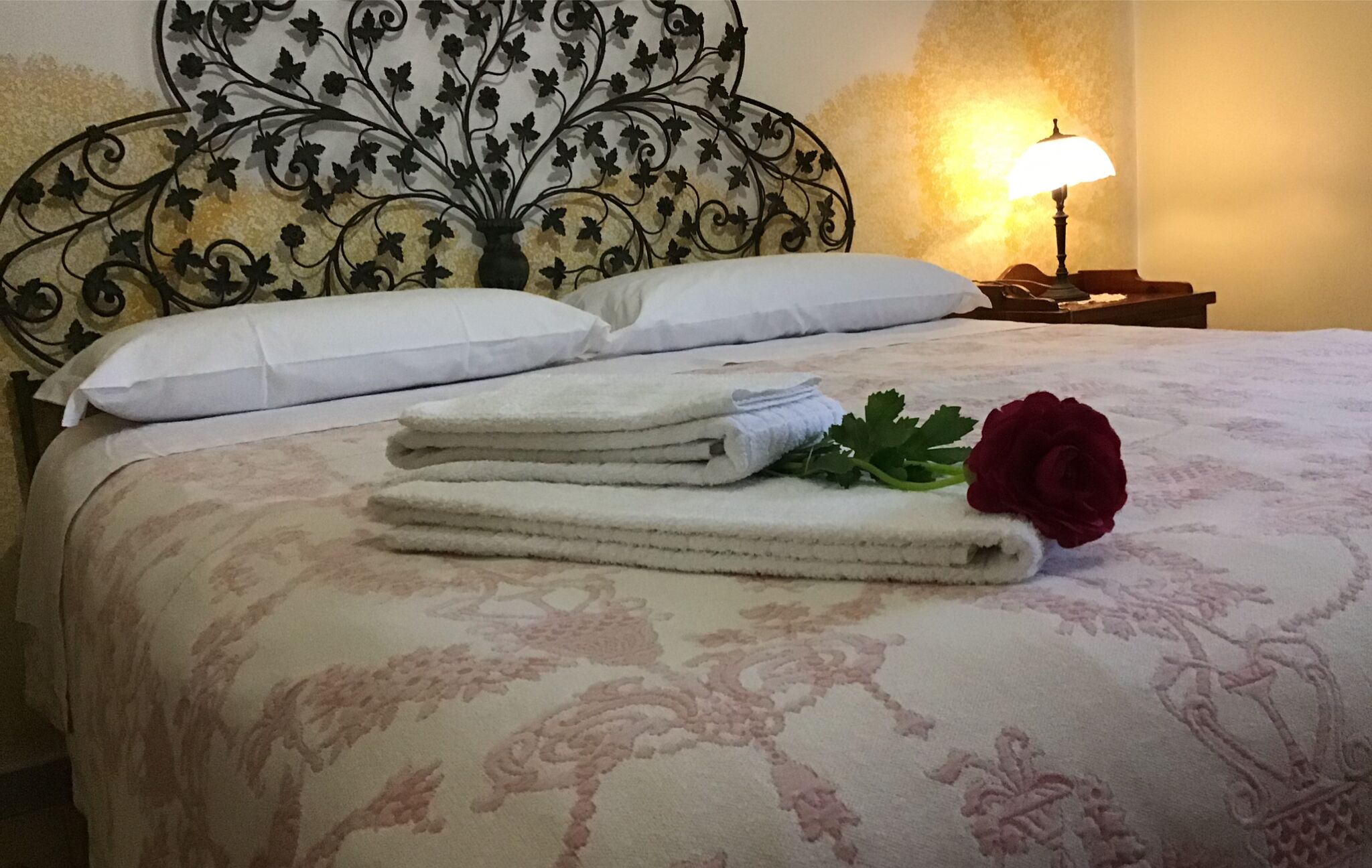 A neatly made bed with a floral-patterned bedspread, a stack of folded towels, a single red rose, and a bedside lamp.