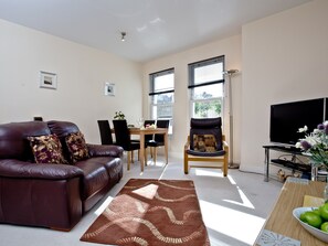 A living room with a leather sofa, dining table, chairs, a TV, and a window with blinds. - A modern well-appointed apartment in a convenient location. (Torquay)