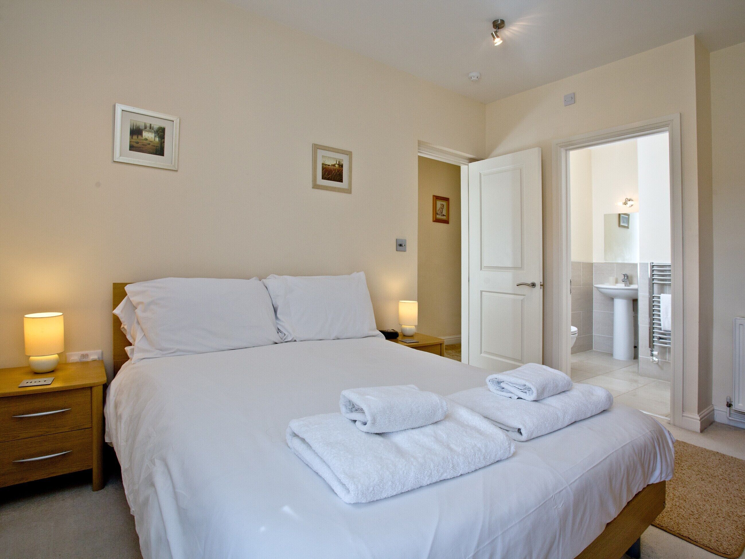 Une chambre d’hôtel comprenant un lit, deux tables de chevet, une commode, une salle de bain avec un lavabo et un miroir, et deux tableaux encadrés accrochés au mur.