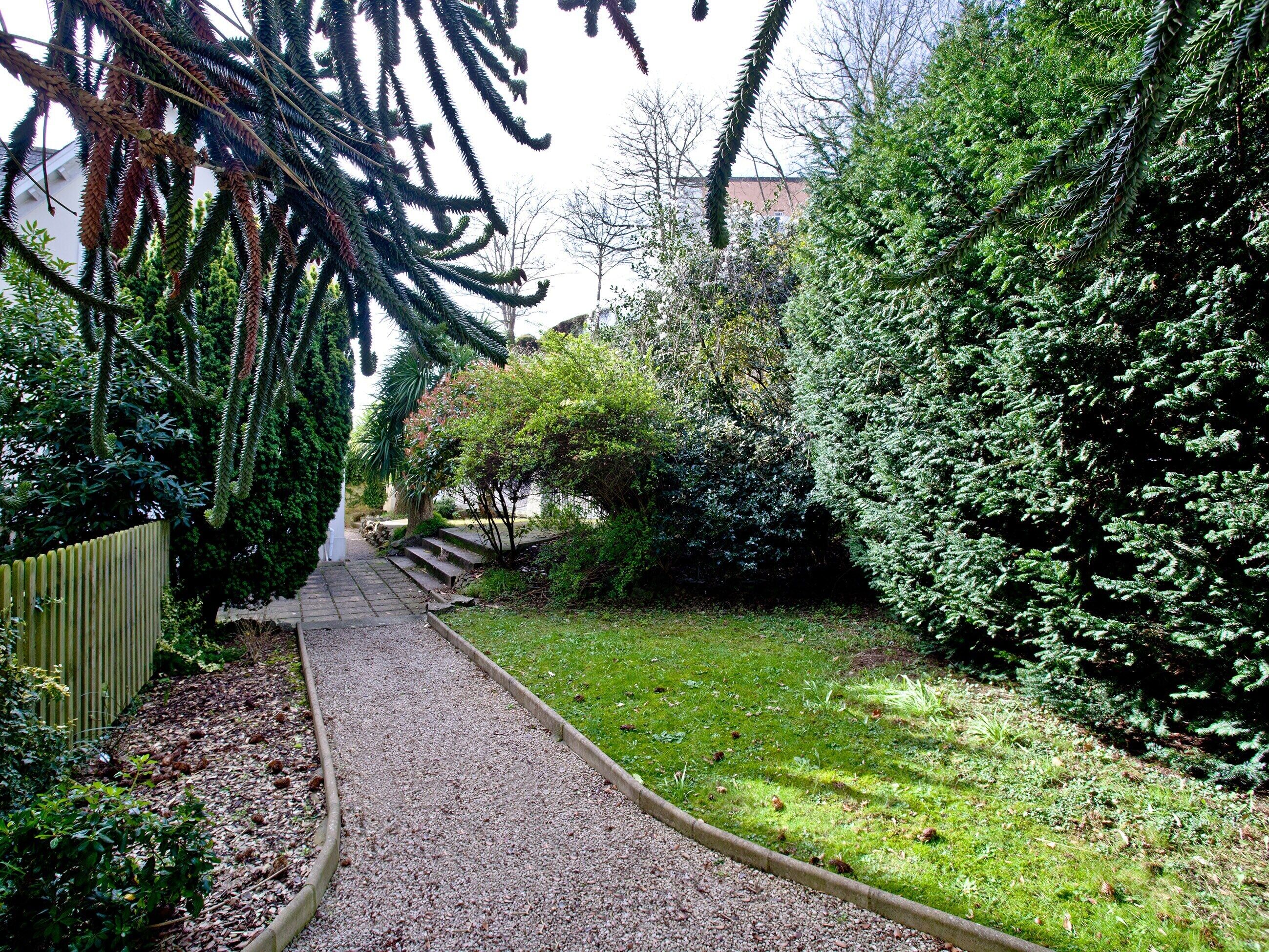 Un chemin de gravier traverse un jardin où la végétation est luxuriante, et une clôture en bois borde le sentier d’un côté.