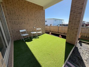Un patio con césped artificial, dos sillas blancas y una valla de madera.