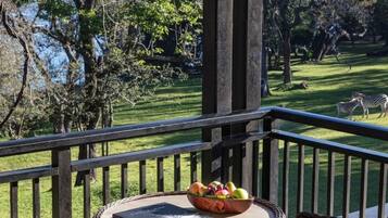 Ein Balkon mit Korbmöbeln, einem Tisch mit Obstschale und Buch, und Blick auf Zebras in einem Park.