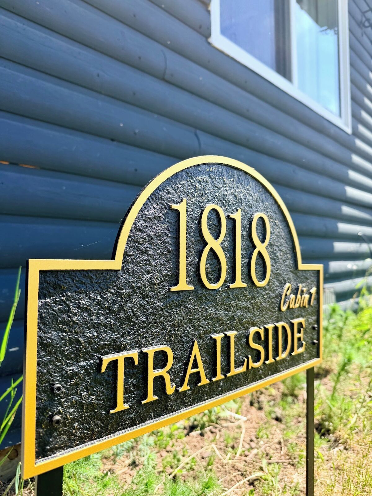 Placa preta e dourada com o número 1818 e as palavras 'TRAILSIDE' e 'Cabana 1', indicando a localização de uma cabana.