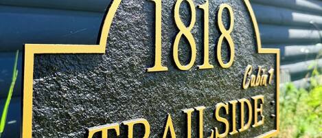 Placa preta e dourada com o número 1818 e as palavras 'TRAILSIDE' e 'Cabana 1', indicando a localização de uma cabana.