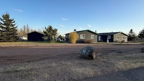A residential area with two houses, a dirt driveway, and a large rock in the foreground. - 1818 Trailside Birch - Cabin 1 (Grand Marais)