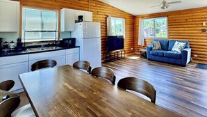 Interior de una cabaña con una mesa de comedor, sillas, una kitchenette, un sofá y un ventilador de techo.