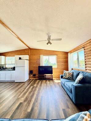 A log cabin interior with a kitchenette, a flat-screen TV, a sofa, and a ceiling fan. - 1818 Trailside Birch - Cabin 1 (Grand Marais)