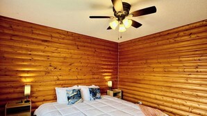 Una habitación con cama, paredes de madera, ventilador de techo y dos lámparas de noche.
