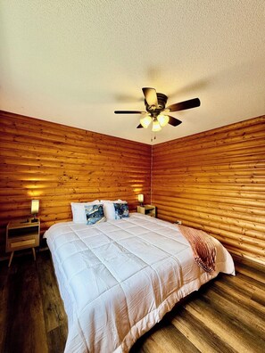 A room with a bed, wooden walls, a ceiling fan, and two bedside lamps. - 1818 Trailside Birch - Cabin 1 (Grand Marais)