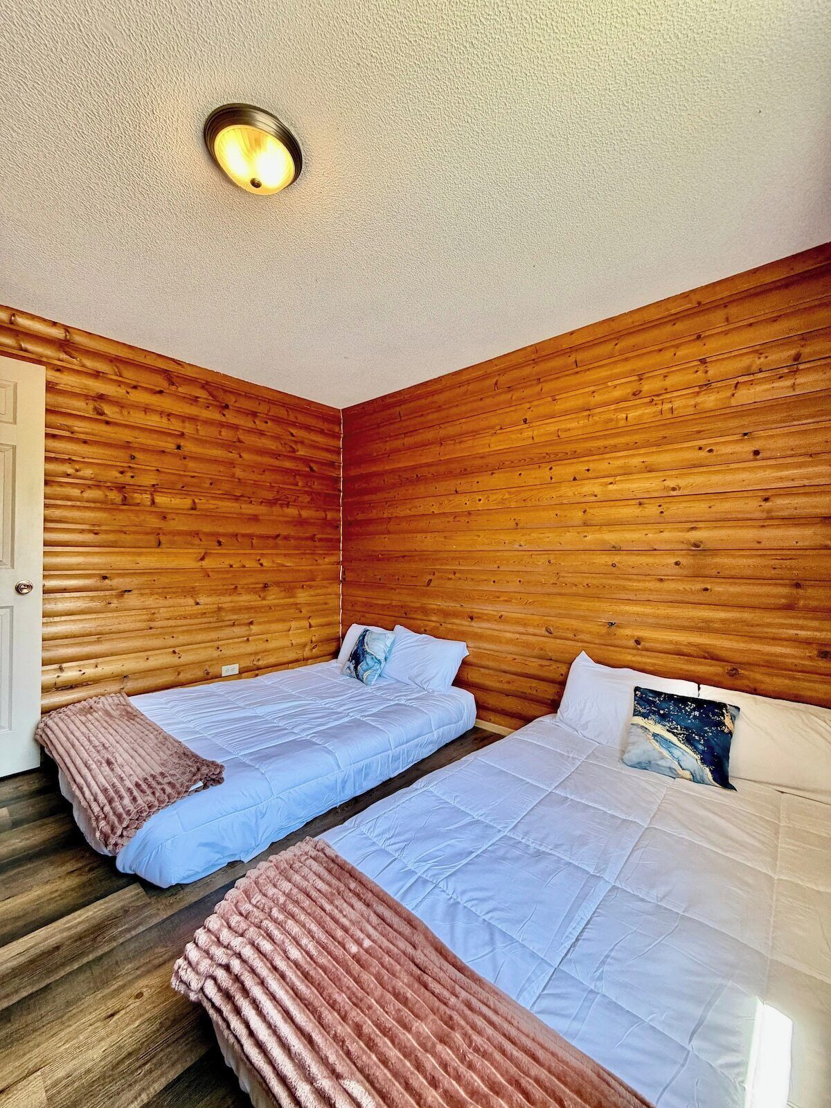 A room with two beds, wooden walls, a ceiling light, and a door.