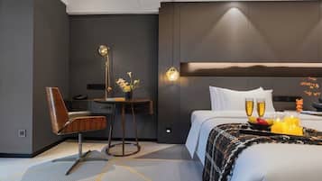 A modern hotel room with a bed, bedside table, chair, and a small desk with a lamp.
