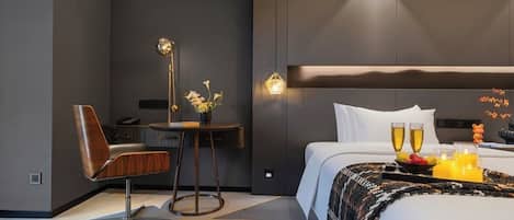 A modern hotel room with a bed, bedside table, chair, and a small desk with a lamp.