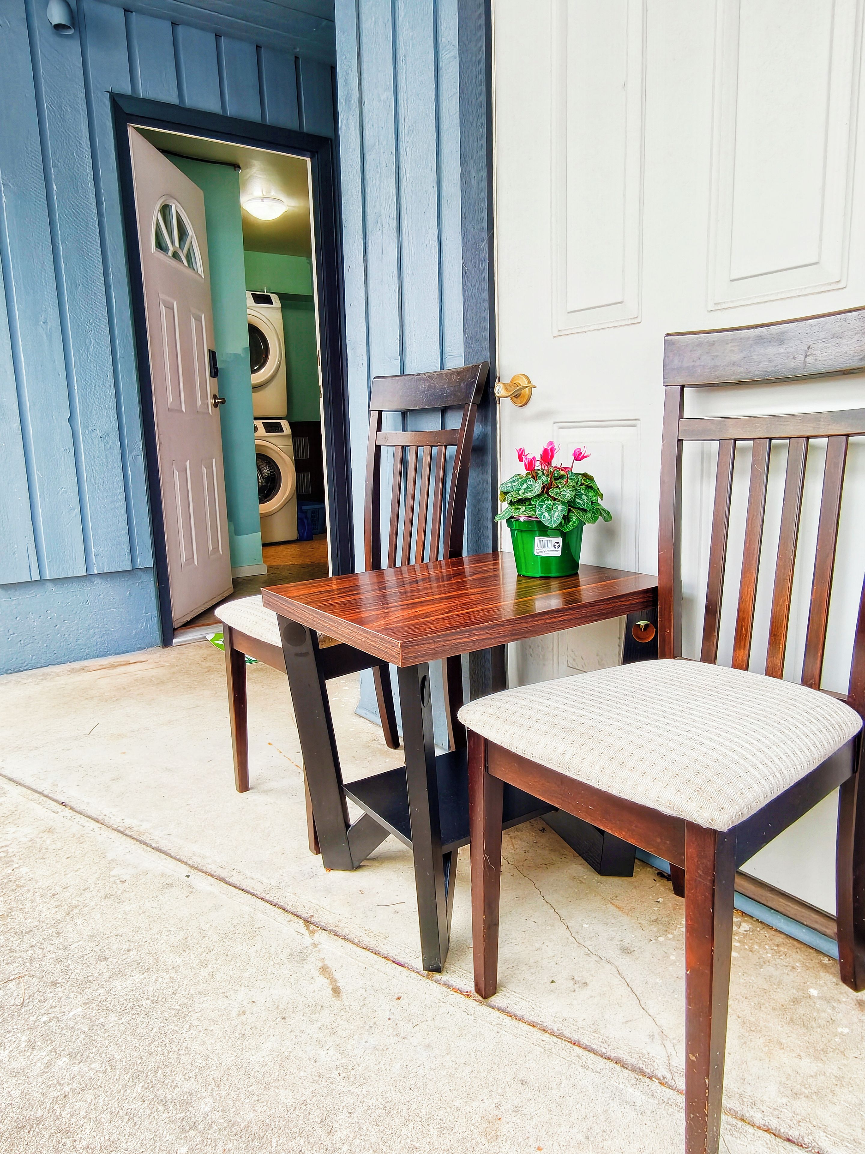 Une table et des chaises en bois se trouvent à l’extérieur d’un bâtiment, près d’une porte qui donne sur une buanderie.
