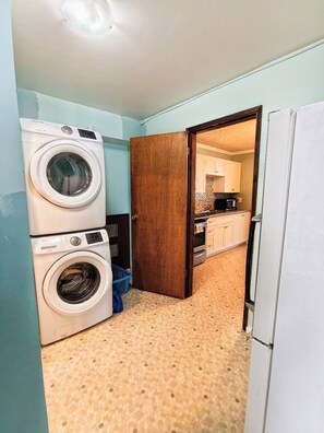 A laundry room with two stacked washers and dryers, a blue laundry basket, and a wooden door leading to a kitchen area.