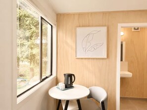 Een kleine kamer met een raam, een lijstwerk, een tafel met een waterkoker en een stoel.