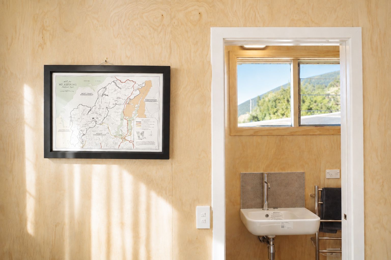 Une carte encadrée du parc national du mont Aspiring est accrochée à un mur en bois, une fenêtre offre une vue sur la verdure, et la salle de bain est équipée d’un lavabo et d’un porte-serviettes.