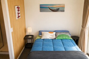 A hotel room with a bed, a nightstand with a lamp, a painting of a boat on the wall, and a view of the sea.