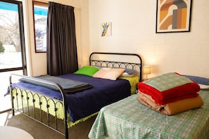 Un lit simple avec un cadre en métal, une fenêtre avec des rideaux et un mur orné d’un tableau.