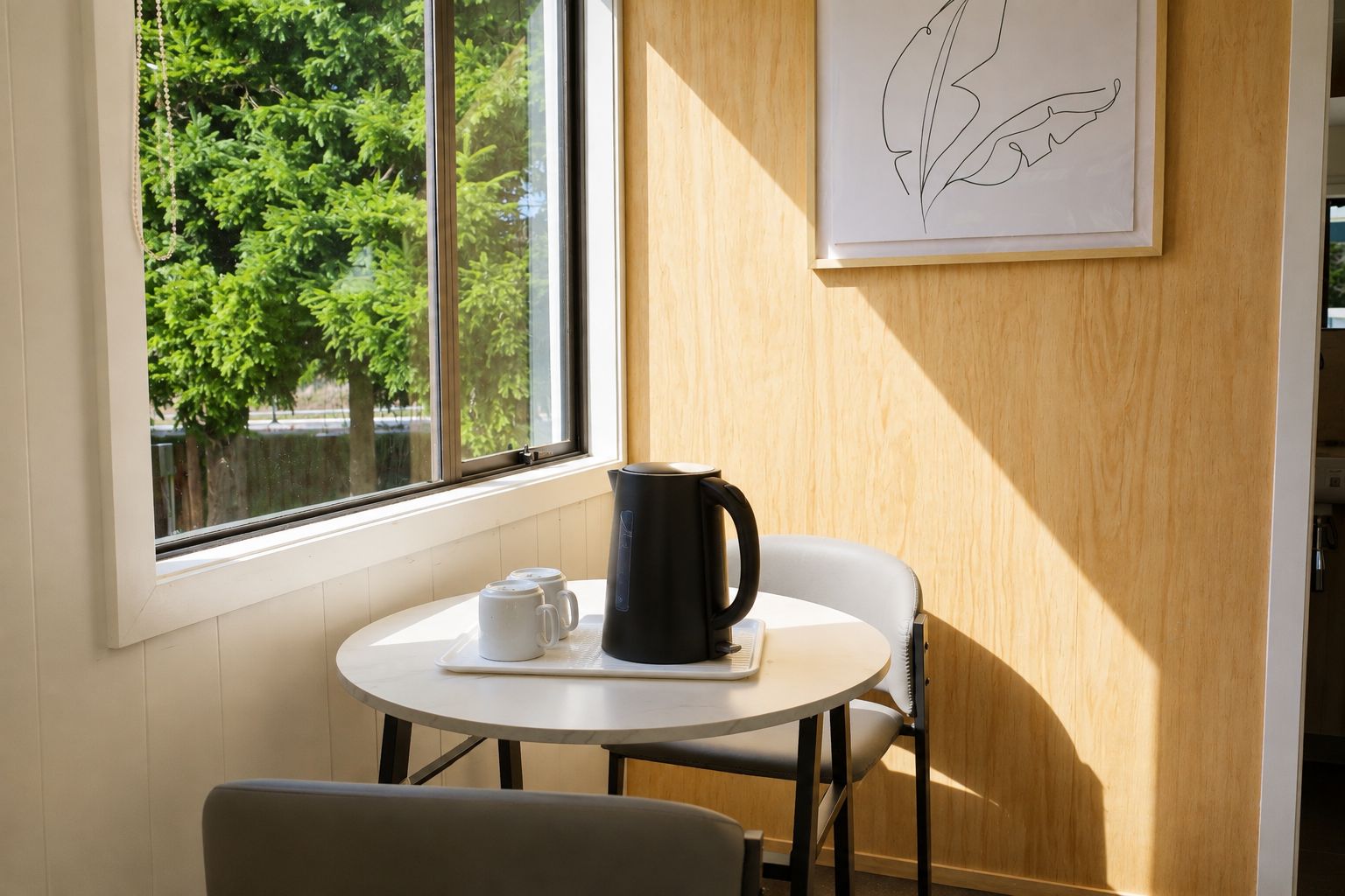 A small round table with a black electric kettle and two white mugs, positioned near a window with a view of trees.