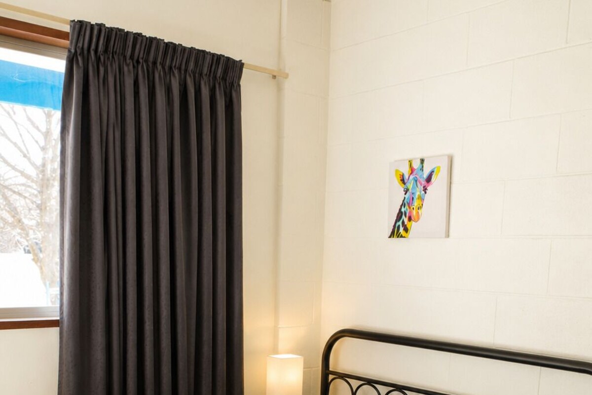 Un lit simple avec un cadre en métal, une couvre-lit bleue et verte et un oreiller. Il y a une fenêtre avec un rideau noir, une lampe fixée au mur et un tableau coloré.