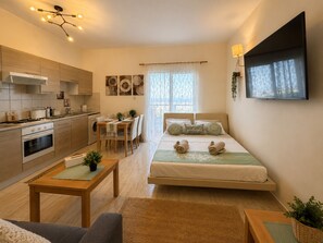 En kompakt bolig med tekøkken, spiseplads, seng med tv og stort vindue.