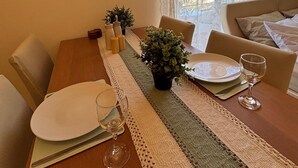 Uma área de refeições com uma mesa de madeira posta para dois, uma planta em vaso e vista para uma varanda através de cortinas leves.