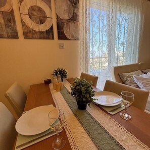 Ein Essbereich mit einem Holztisch, gedeckt für zwei Personen, einer Topfpflanze und Blick auf einen Balkon durch transparente Vorhänge.