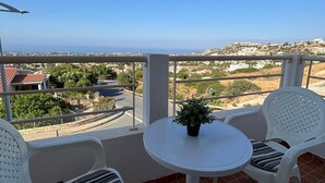 Uma varanda com uma mesa branca e duas cadeiras, um vaso com planta e vista para uma cidade costeira.