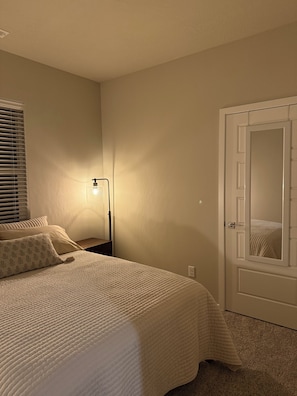 Ein Schlafzimmer mit Bett, Nachttisch, Lampe, einer Tür und einem Fenster mit Jalousien.