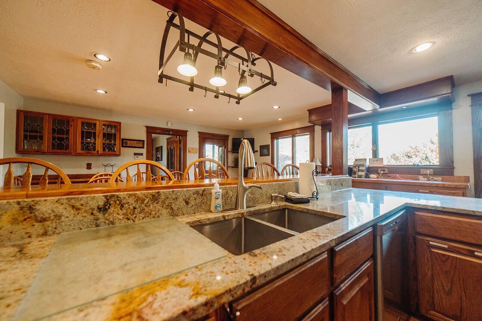 A kitchen with granite countertops, a stainless steel sink, wooden chairs, and a large window providing natural light.