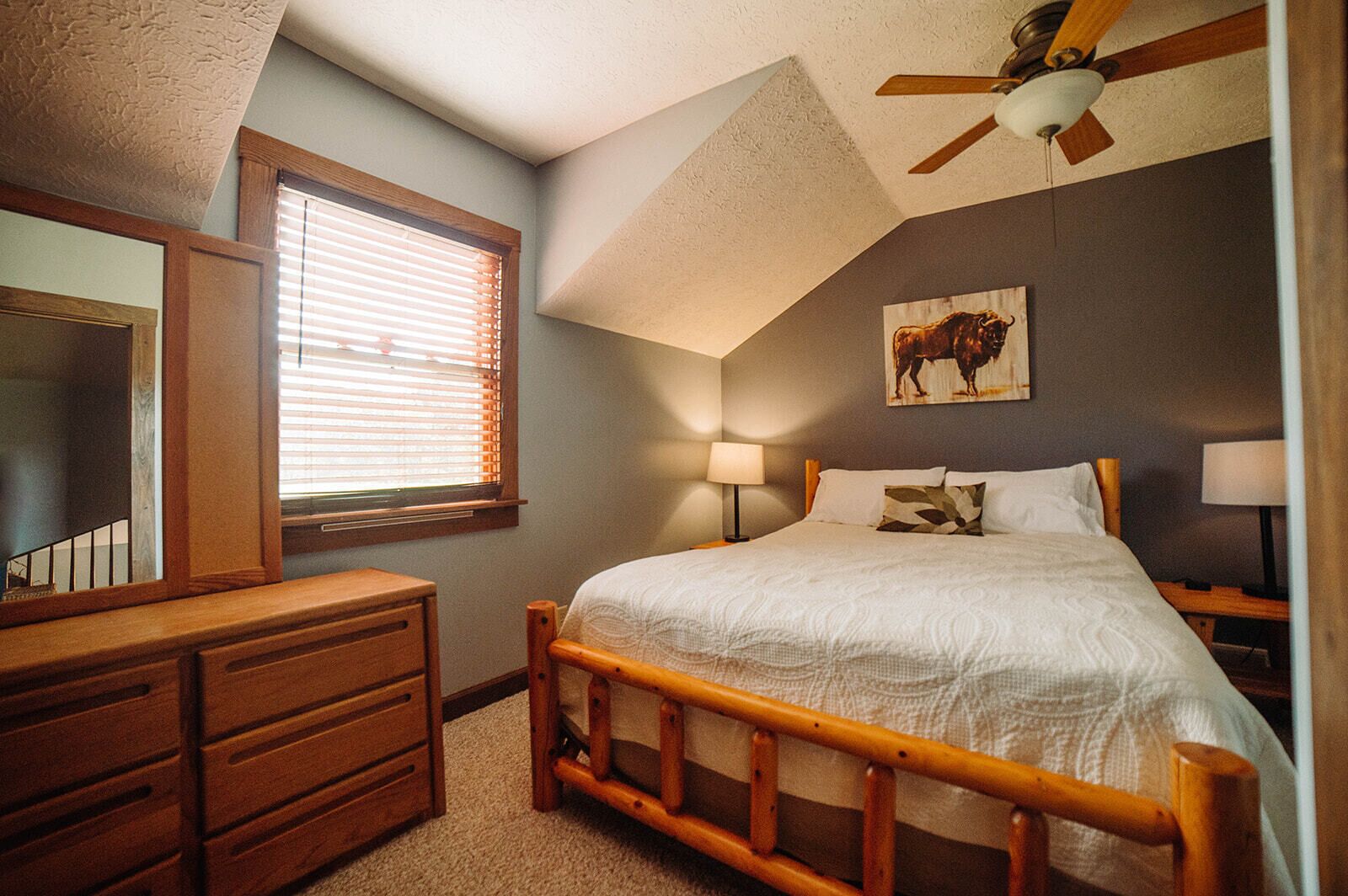 A bedroom with a wooden bed, a dresser, a ceiling fan, a window with blinds, and a wall-mounted lamp.